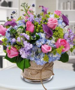 A stunning bouquet of calla lilies in purple, pink roses, hydrangea and elements like thistle to add texture. A lovely lace ribbon with raffia is wrapped around the vase combining modern and vintage.