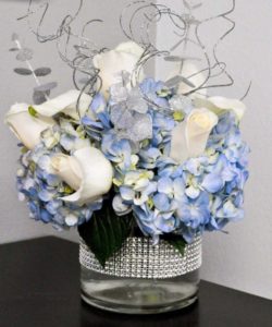 modern arrangement features light blue hydrangea and premium white calla lilies accented with sparkles, blue gems, silver leaves, and glistening curly willow.